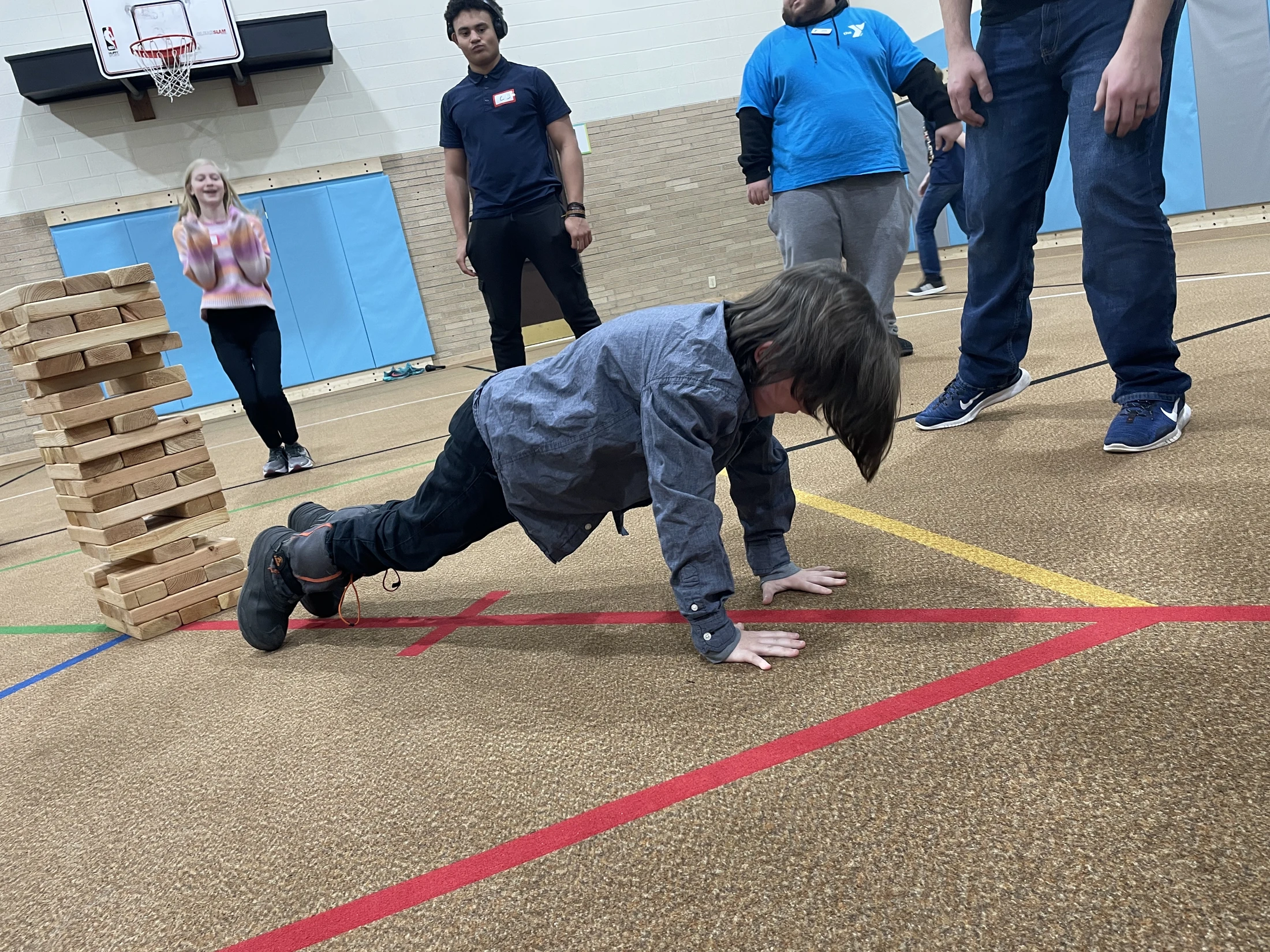 A young boy holds a plank position on a gym floor next to a tall, unstable tower of giant wooden blocks as other children and adults watch.