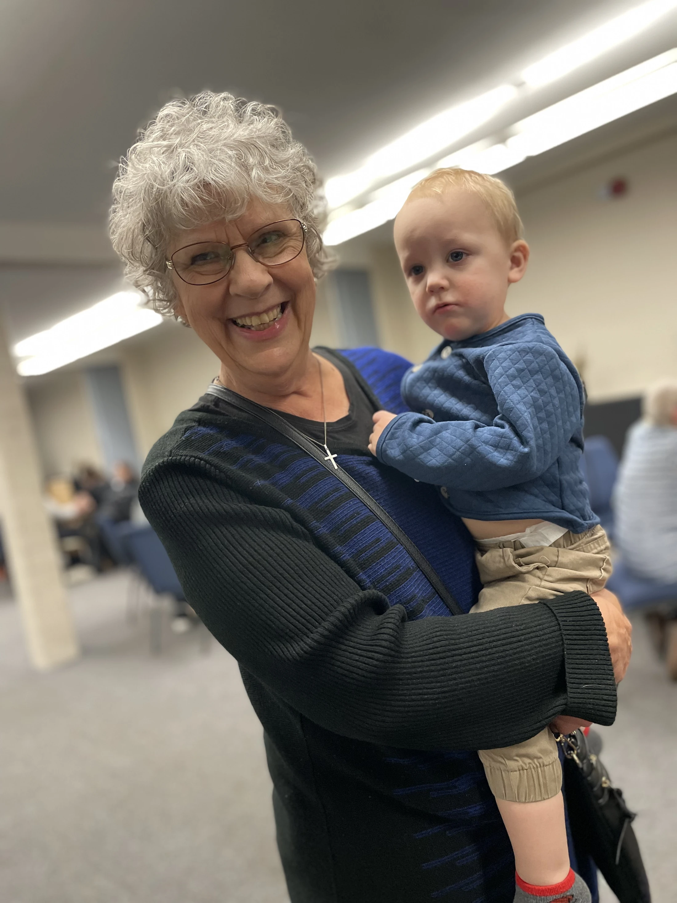A happy older woman with short grey hair and glasses smiles while holding a toddler who has a grumpy expression. They are indoors in a large room.