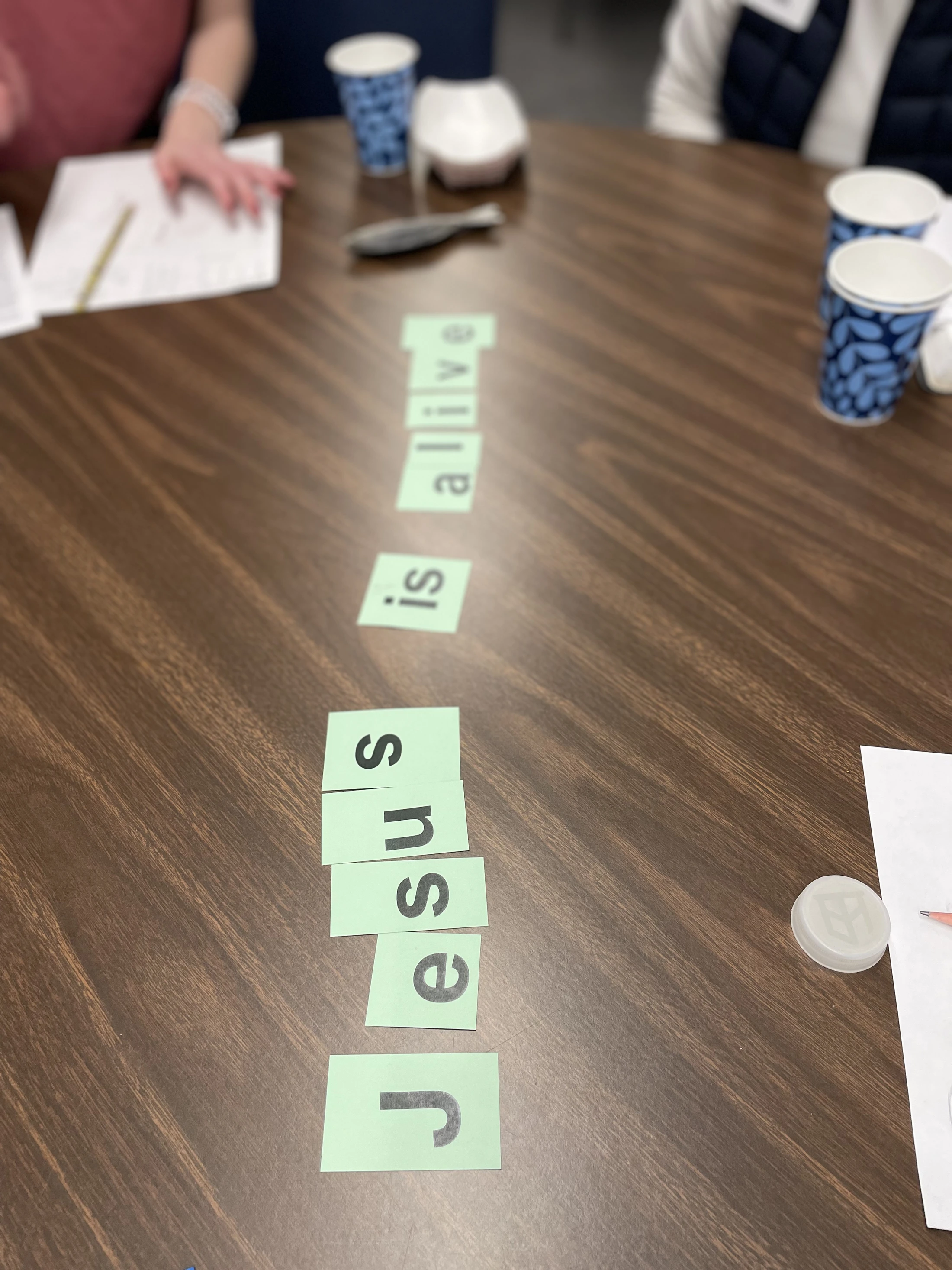 Green paper cards with letters spelling out 'Jesus is alive' arranged vertically on a dark brown wooden table during a group activity.