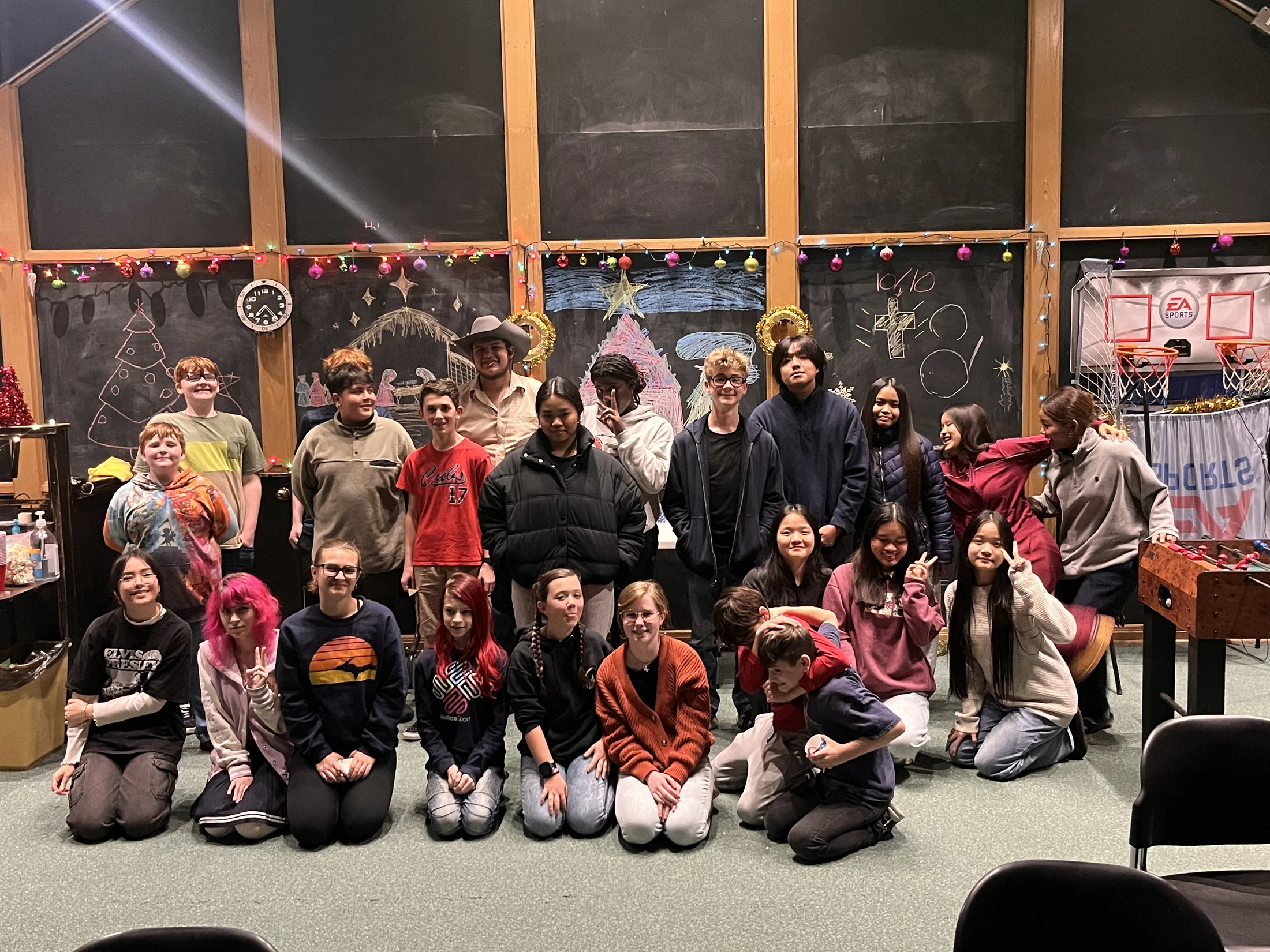 A large, diverse group of young people posing for a photo in a classroom decorated for Christmas, with chalk drawings on blackboards behind them.