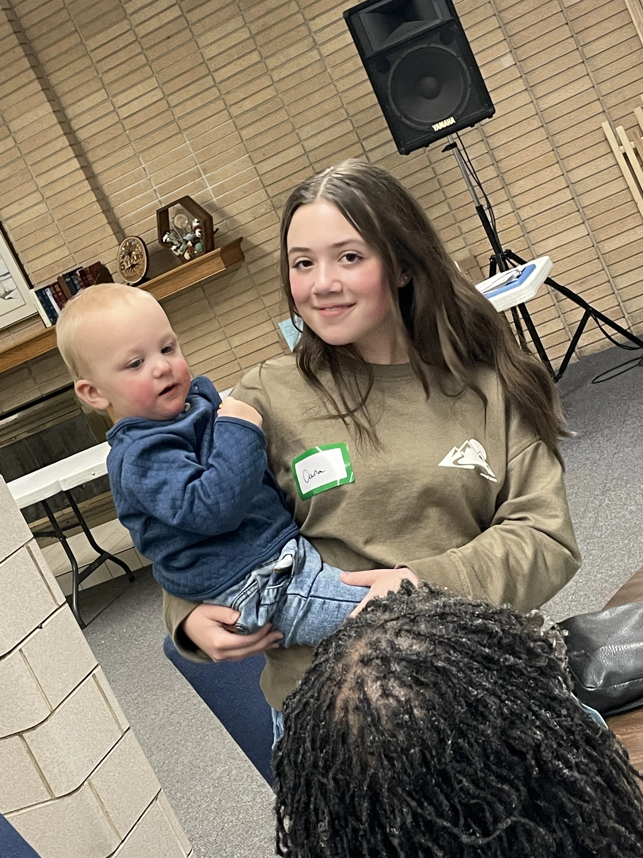 A smiling young woman with long brown hair holds a blond baby with rosy cheeks. They are indoors in a room with a tan brick wall.