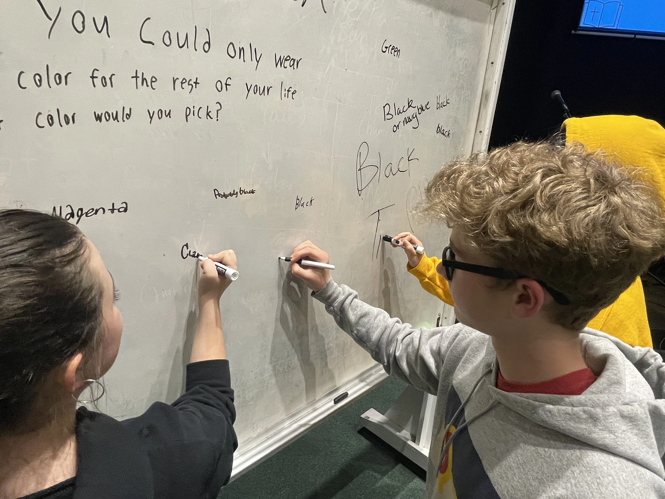 Several young people write their answers with markers on a whiteboard that asks what single color they would wear for the rest of their lives.
