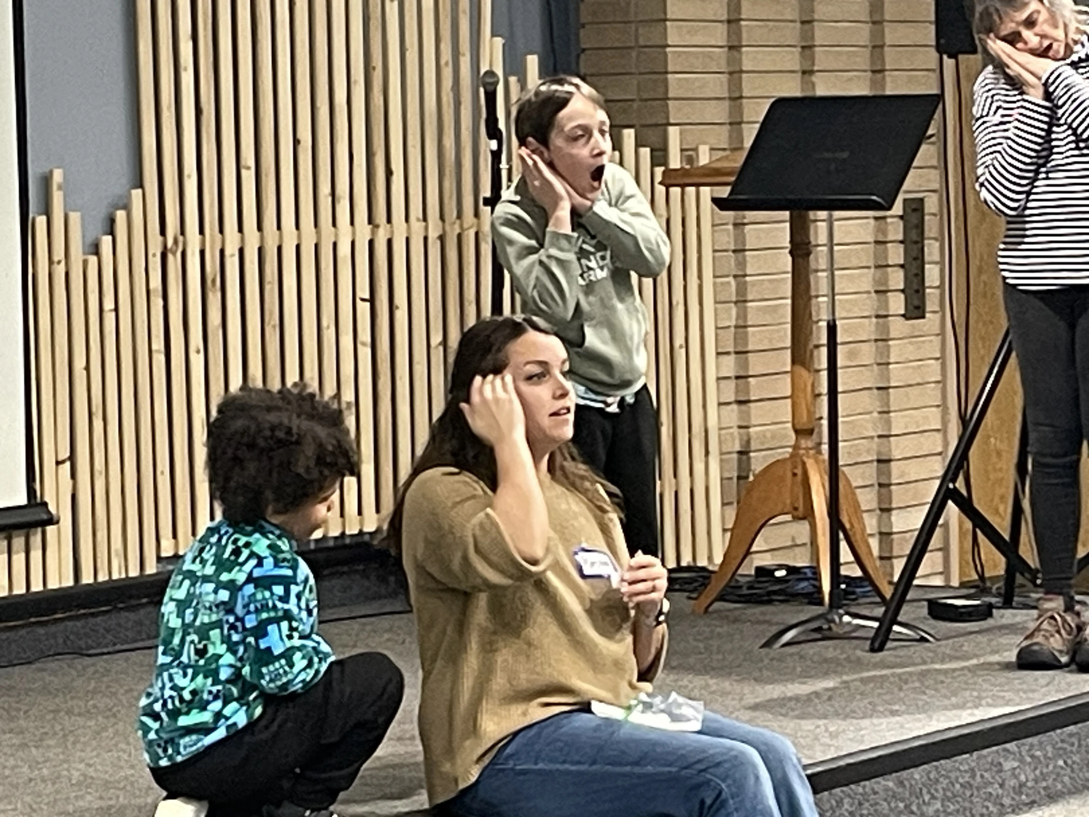 A group of people on a stage during an activity. A child stands with a shocked expression, a woman mimes sleeping, and another woman and child watch intently.