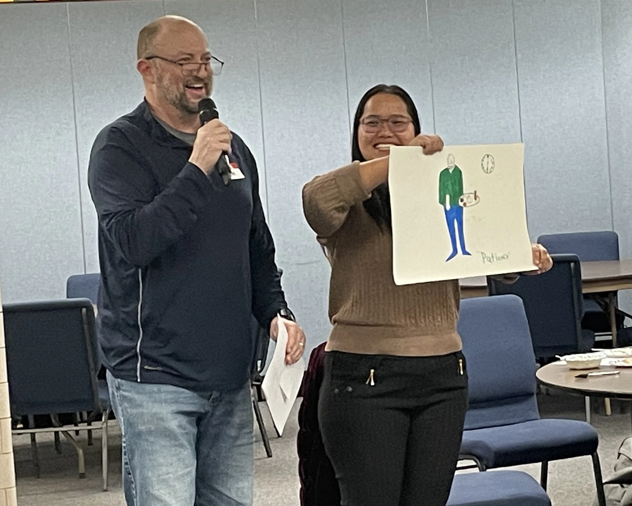 A smiling man with a microphone stands next to a smiling woman who proudly holds up a colorful drawing of a person.