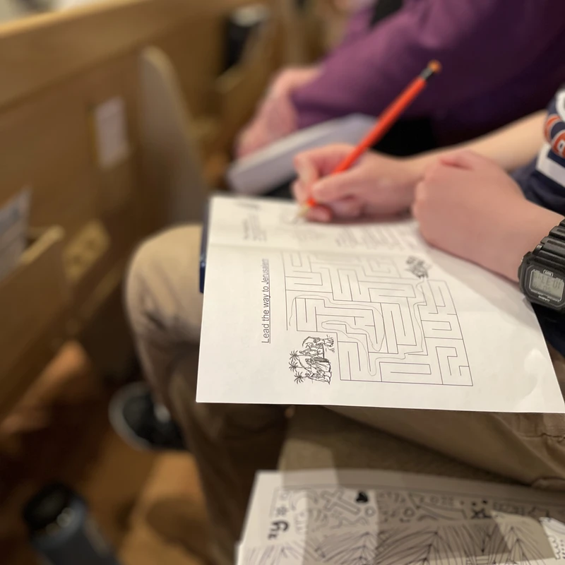 A child sits in a pew using an orange pencil to complete a printed maze activity sheet titled 'Lead the way to Jerusalem' during a service.