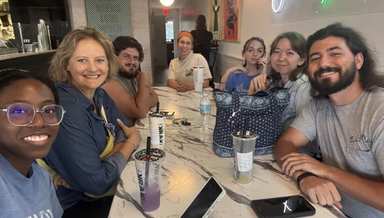 A diverse group of seven young adults sit around a long marble table in a cafe, smiling at the camera with their bubble tea drinks in front of them.