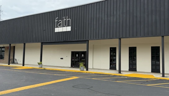 The storefront of a modern building with a black corrugated metal facade and a white brick lower level. A sign says 'faith' in white lowercase letters.