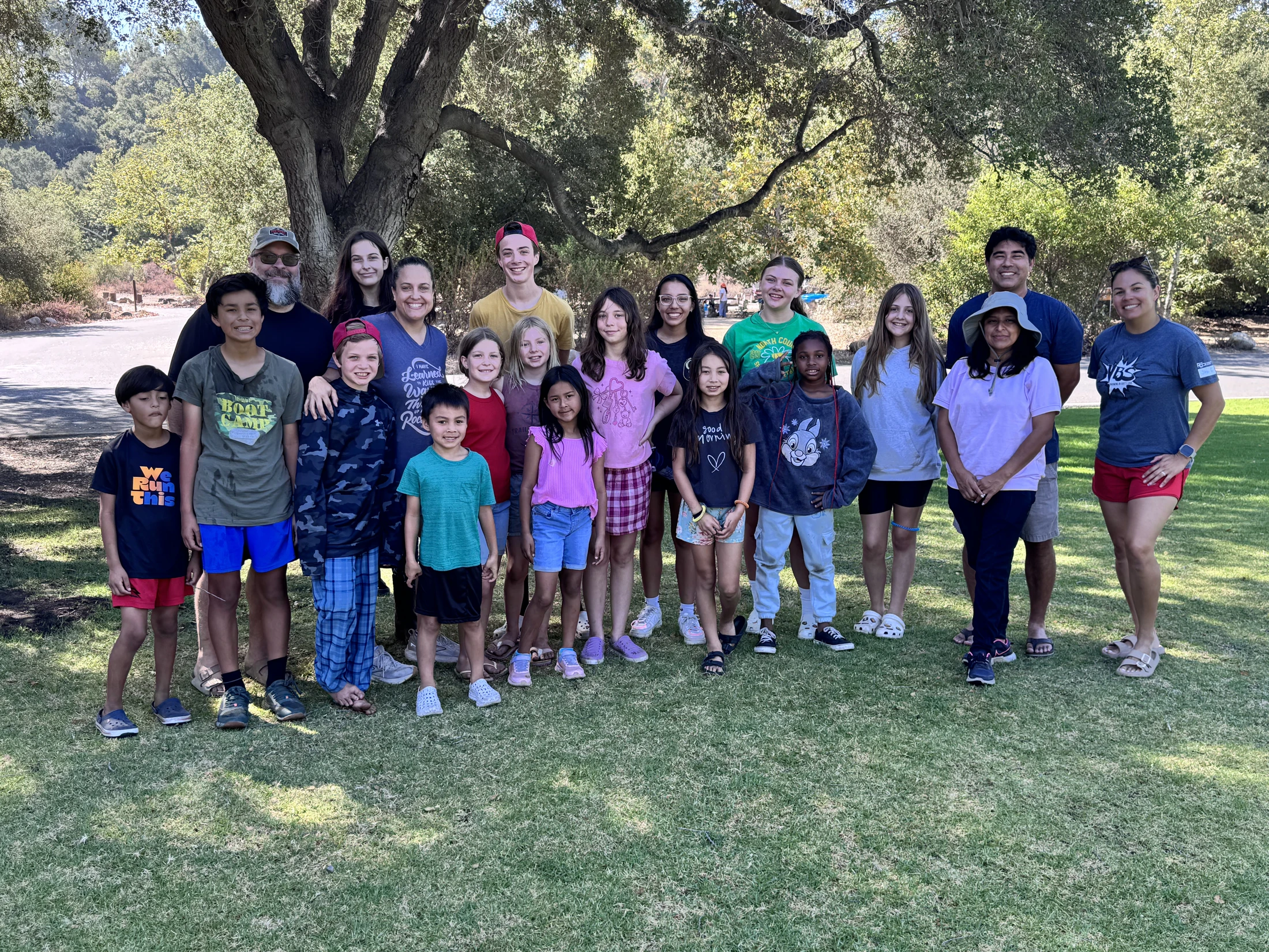 A diverse group of adults and children smiling for a photo on a sunny day. They are standing on a green lawn in front of a large tree and a wooded background.