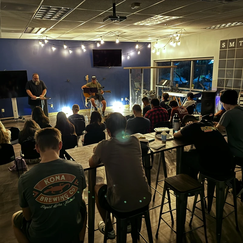 A group of young people seated in a dimly lit room, listening to a man speaking at a podium and another man playing an acoustic guitar.