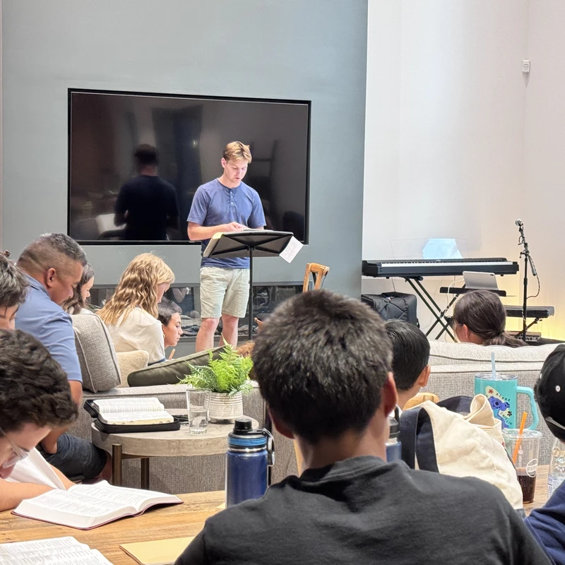 A young man stands at a lectern, reading to a group of people seated on couches in a modern living room during a bible study session.