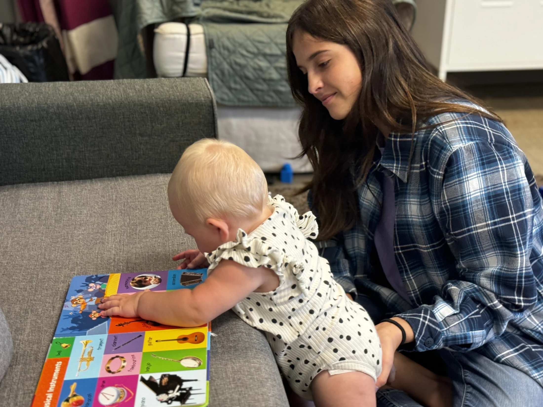 A woman in a plaid shirt sits on a couch and looks lovingly at a blonde baby who is pointing at a colorful board book about musical instruments.