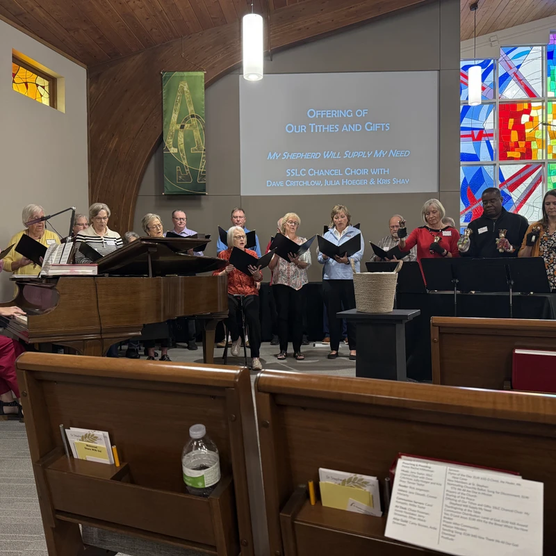A diverse church choir sings from binders, accompanied by a woman playing a grand piano. Behind them is a projection screen and a colorful stained glass window.