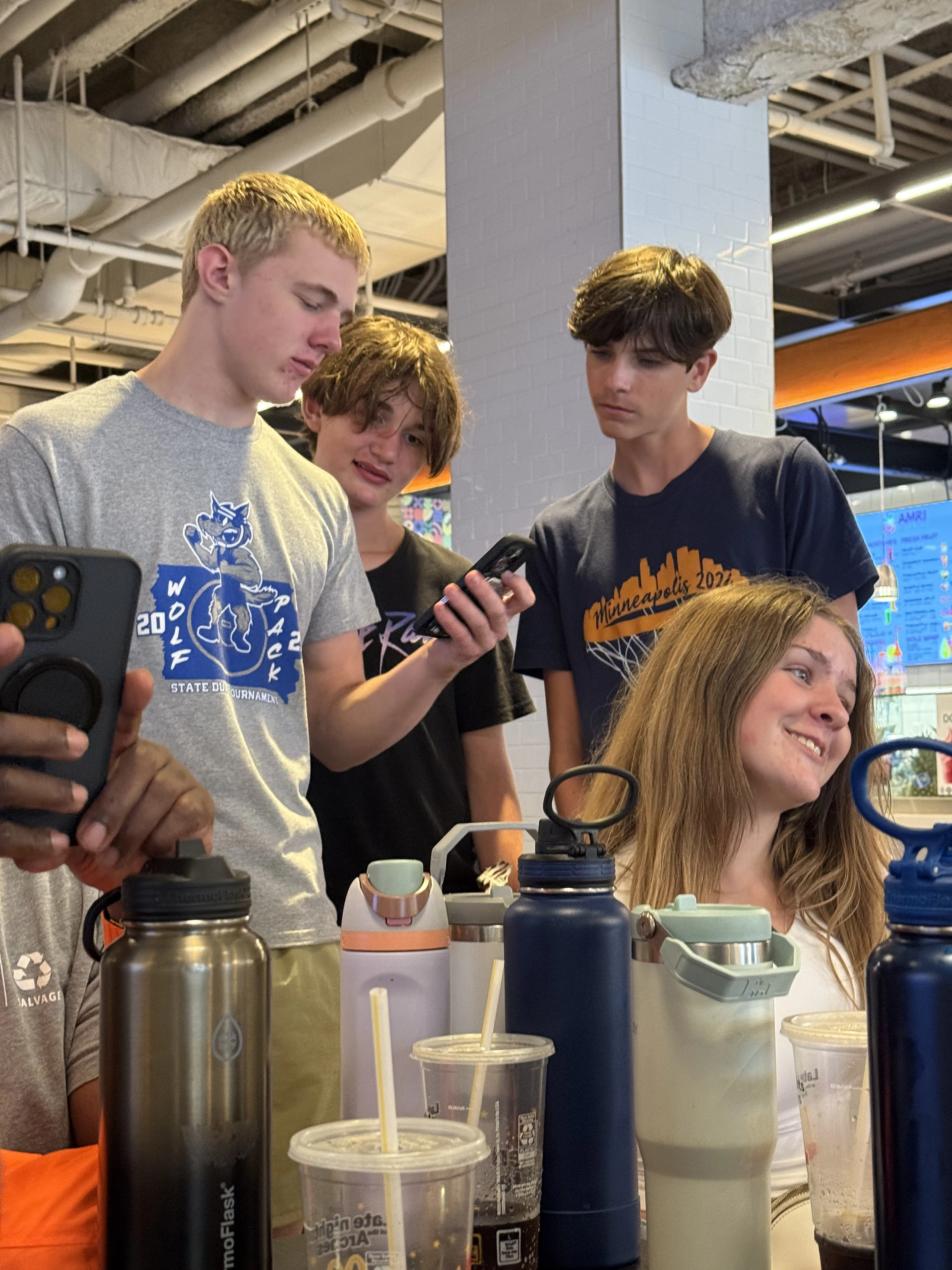 A group of four teenagers gathered indoors. Three boys stand together looking at a phone, while a girl sits at a table in front of them, smiling.