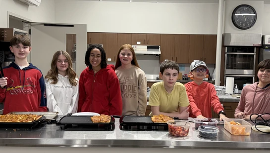 A group of seven young students stand behind a stainless steel counter in a kitchen, posing with the pizzas and fresh fruit they prepared.