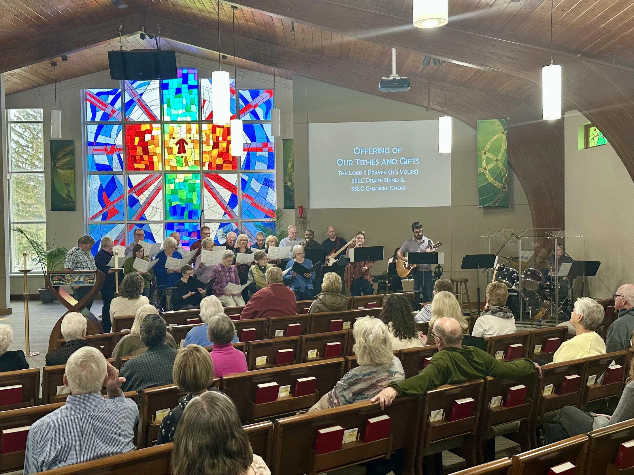 A congregation seated in pews watches a choir and band perform at the front of a church sanctuary in front of a large, colorful stained-glass window.