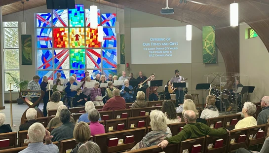 A congregation seated in pews watches a choir and band perform at the front of a church sanctuary in front of a large, colorful stained-glass window.