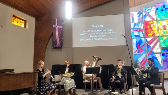 A five-piece brass ensemble performs on stage in a church. The musicians play trumpets, a French horn, a tuba, and a trombone in front of music stands.