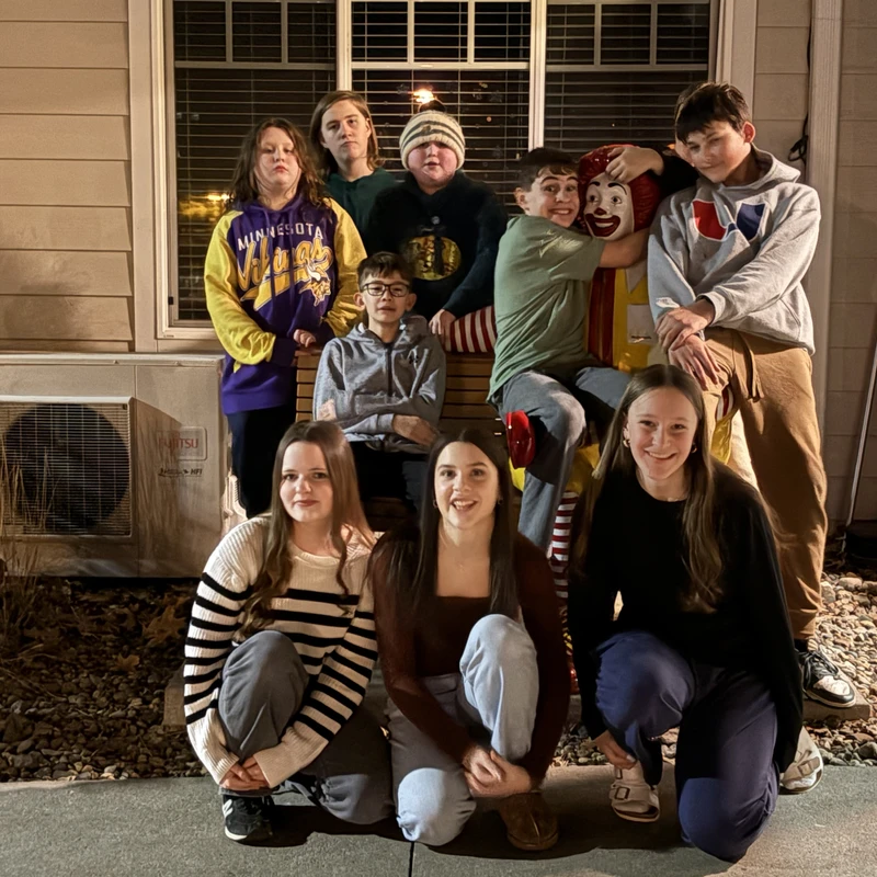 A group of ten young people pose for a picture outdoors at night around a bench, with one boy enthusiastically hugging a Ronald McDonald statue.