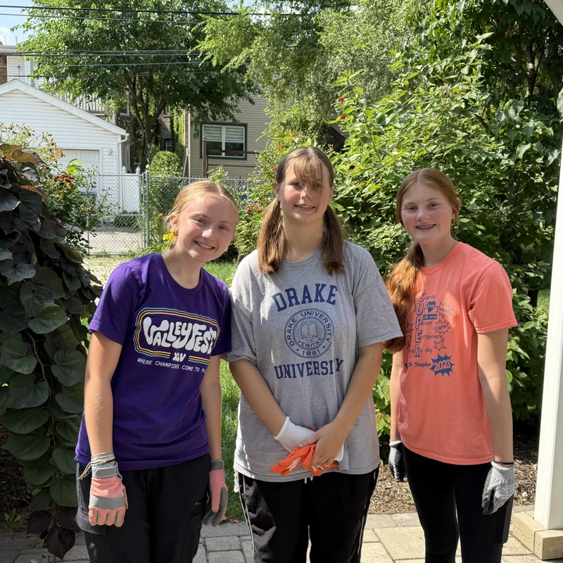 Three smiling teenage girls in casual t-shirts and work gloves stand together in a sunny backyard garden, ready for yard work.
