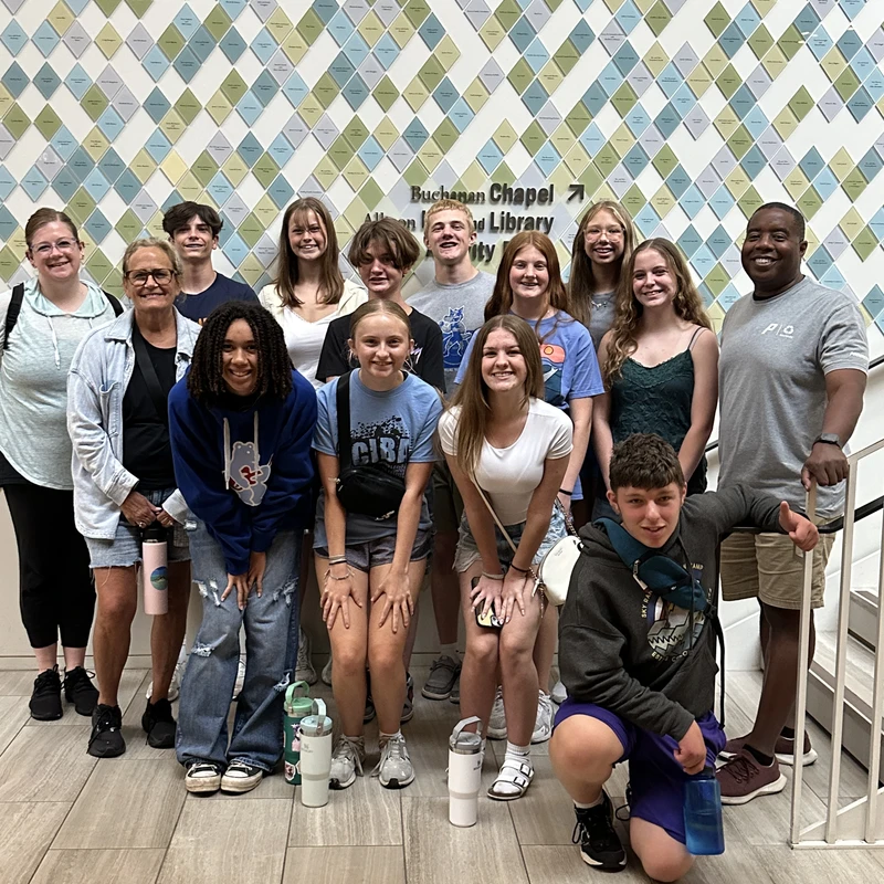 A diverse group of teenagers and adults smile for a picture. They are standing indoors in front of a wall with a geometric pattern of blue and green tiles.
