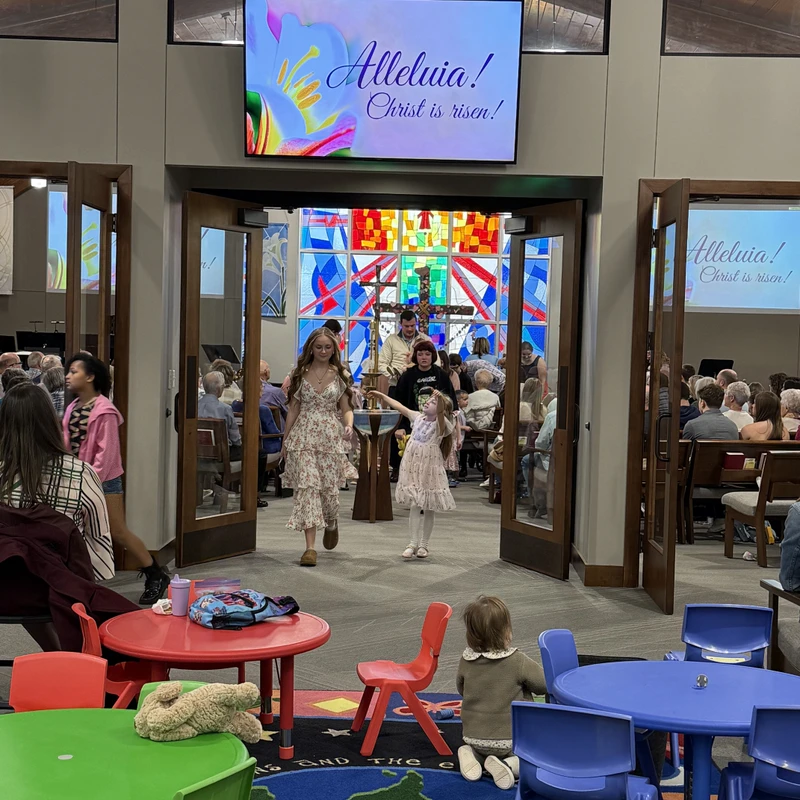 A view from a children's area with colorful tables into a church sanctuary. People exit the service under a screen that says 'Alleluia! Christ is risen!'