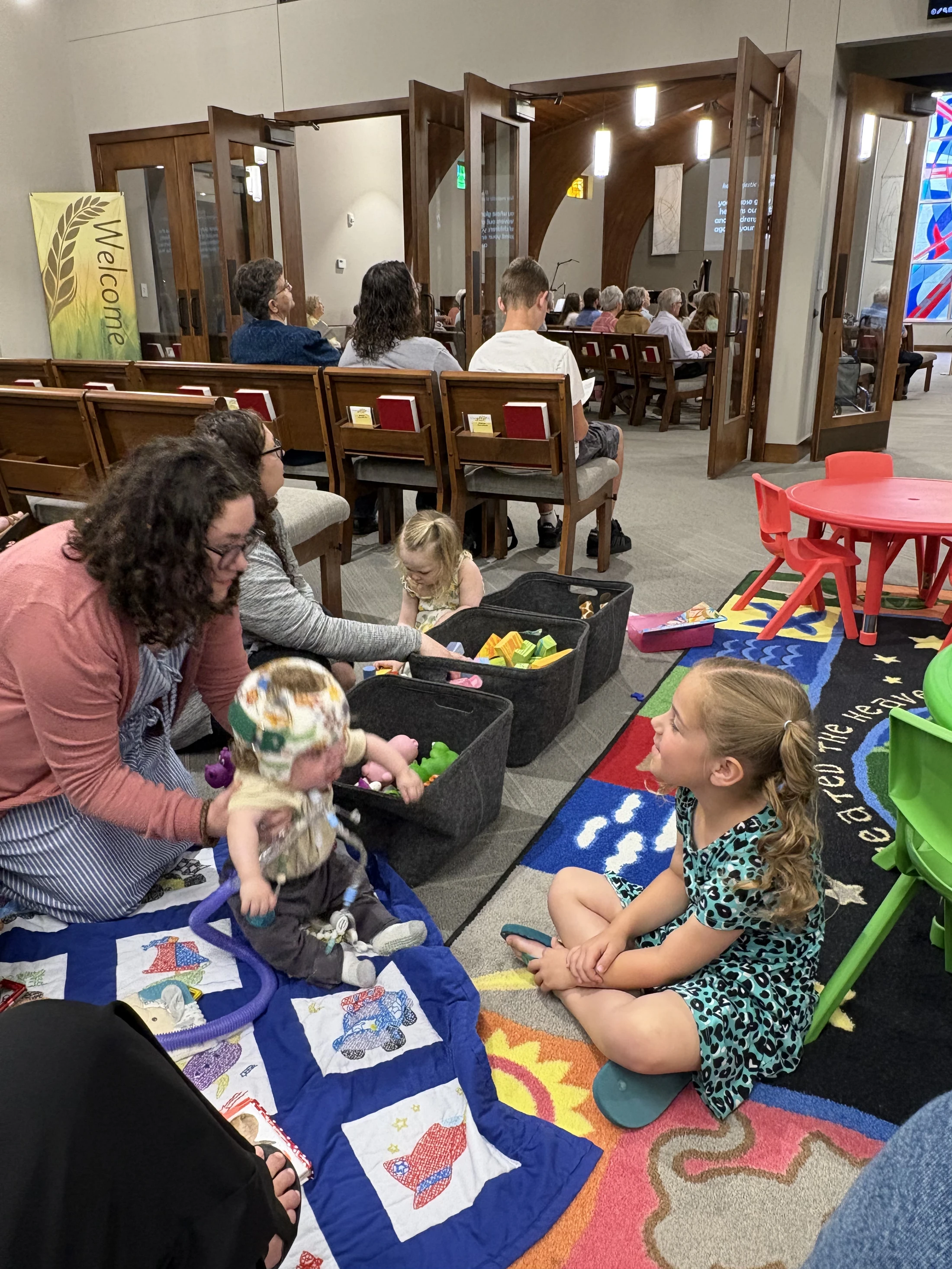 Mothers and young children sit on a colorful rug in a play area at the back of a church, with the congregation seated in pews visible beyond.