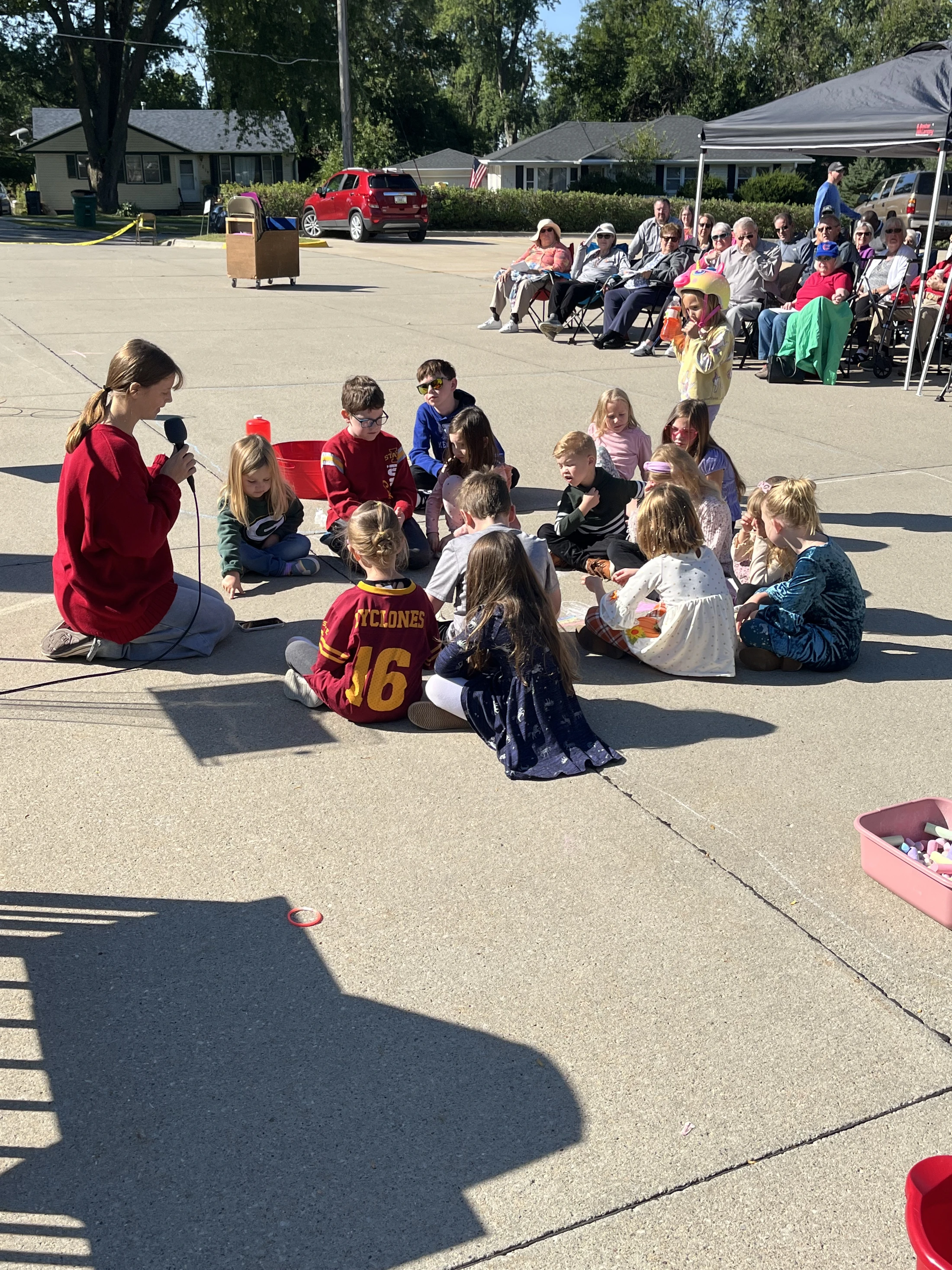 A young woman in a red sweater kneels on pavement, speaking into a microphone to a group of children sitting in a circle. Adults watch from the background.