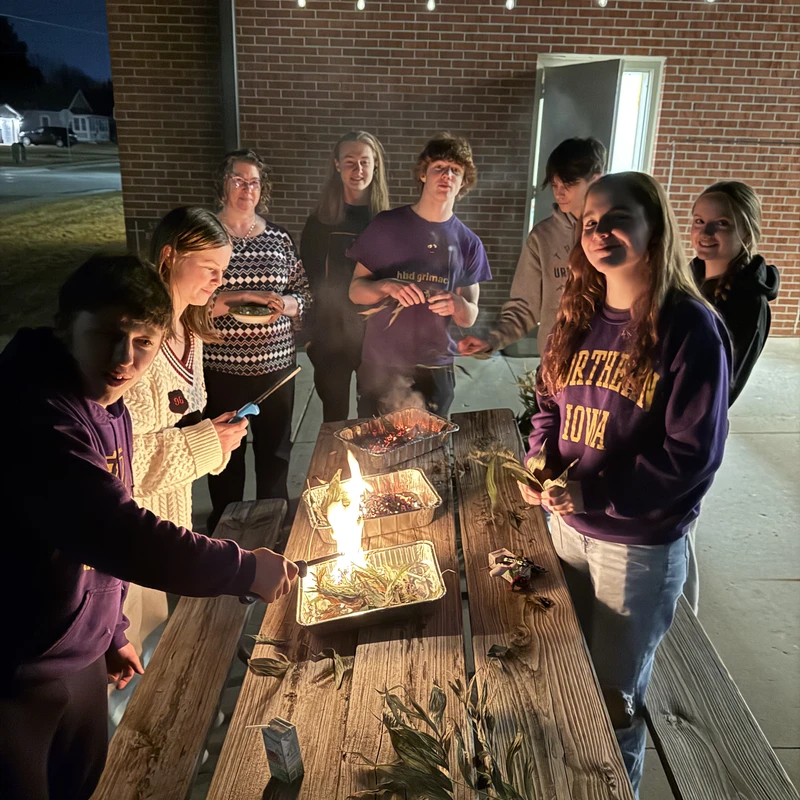 A group of young people and adults stand around a picnic table at night, burning leaves in foil pans under string lights against a brick wall.