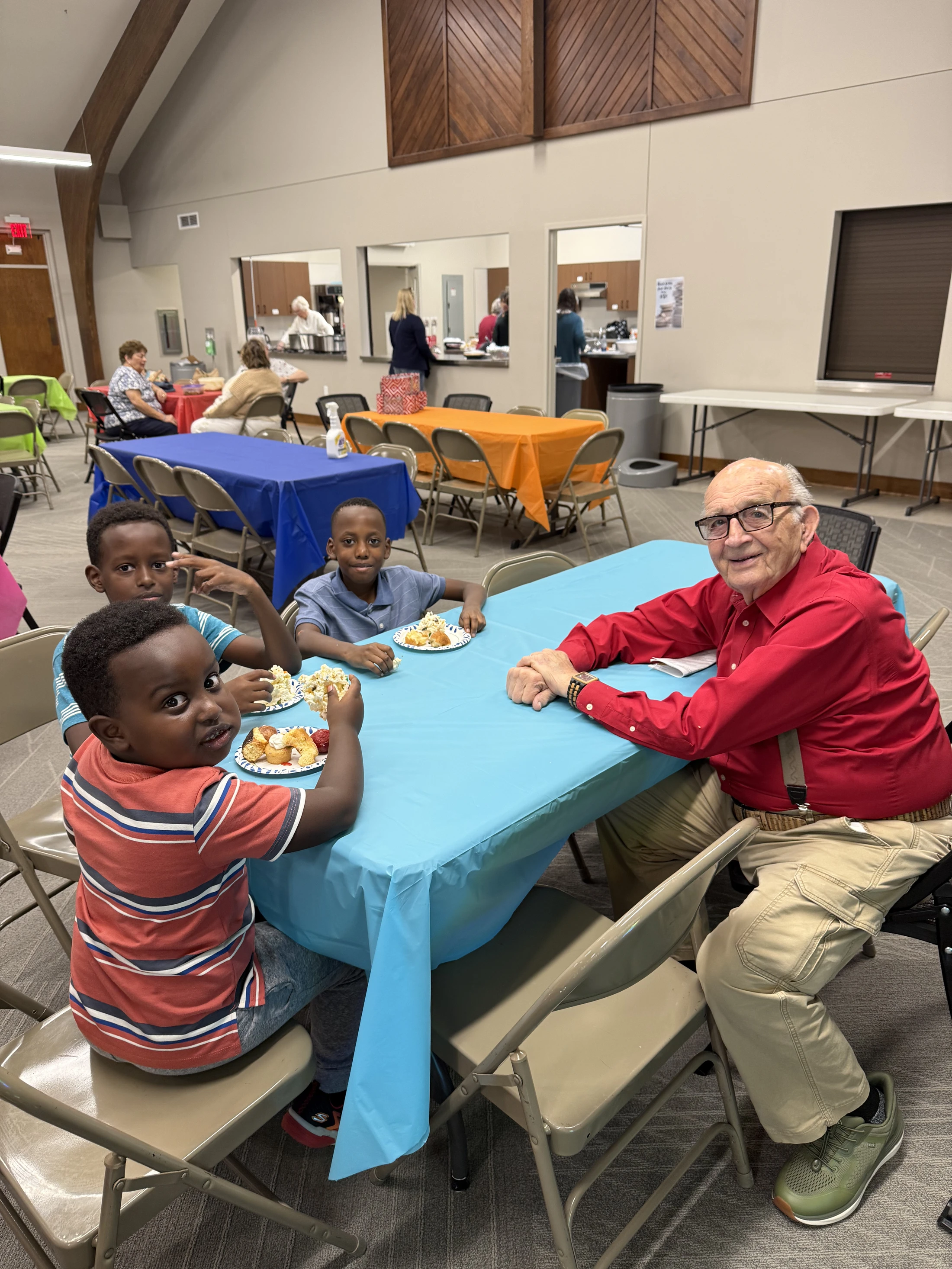 An elderly white man and three young Black boys smile for the camera while sitting together at a table with a blue cloth during a community meal.