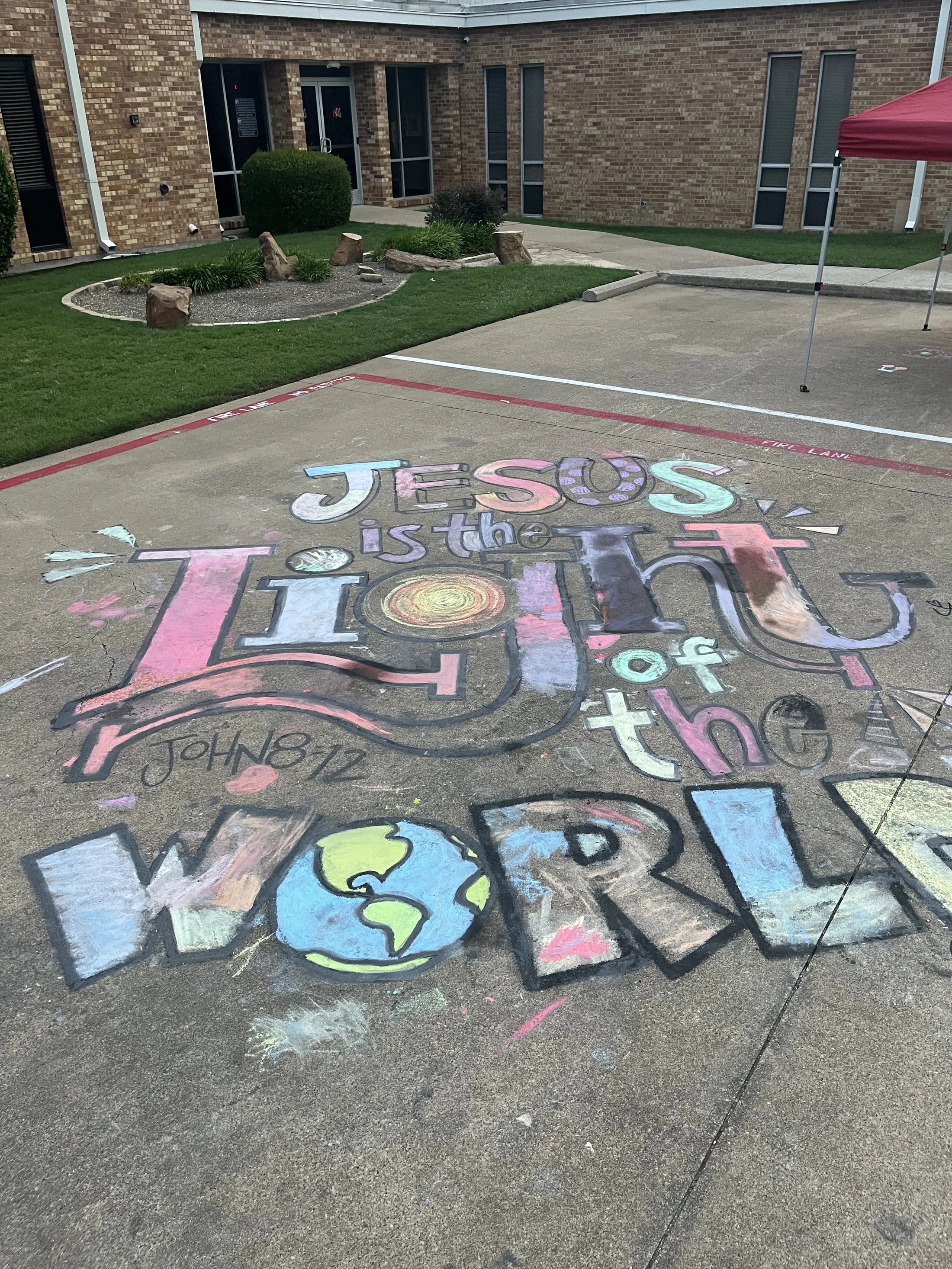 A large, colorful chalk drawing on a concrete pavement that reads, 'Jesus is the Light of the World,' with an illustration of the Earth.