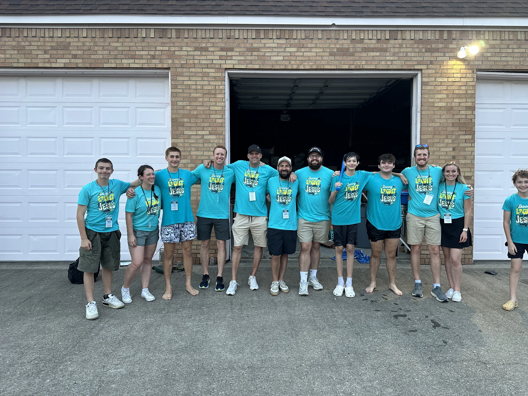 A group of adults and teenagers pose for a photo in front of a brick building with garage doors, all wearing matching bright blue t-shirts.