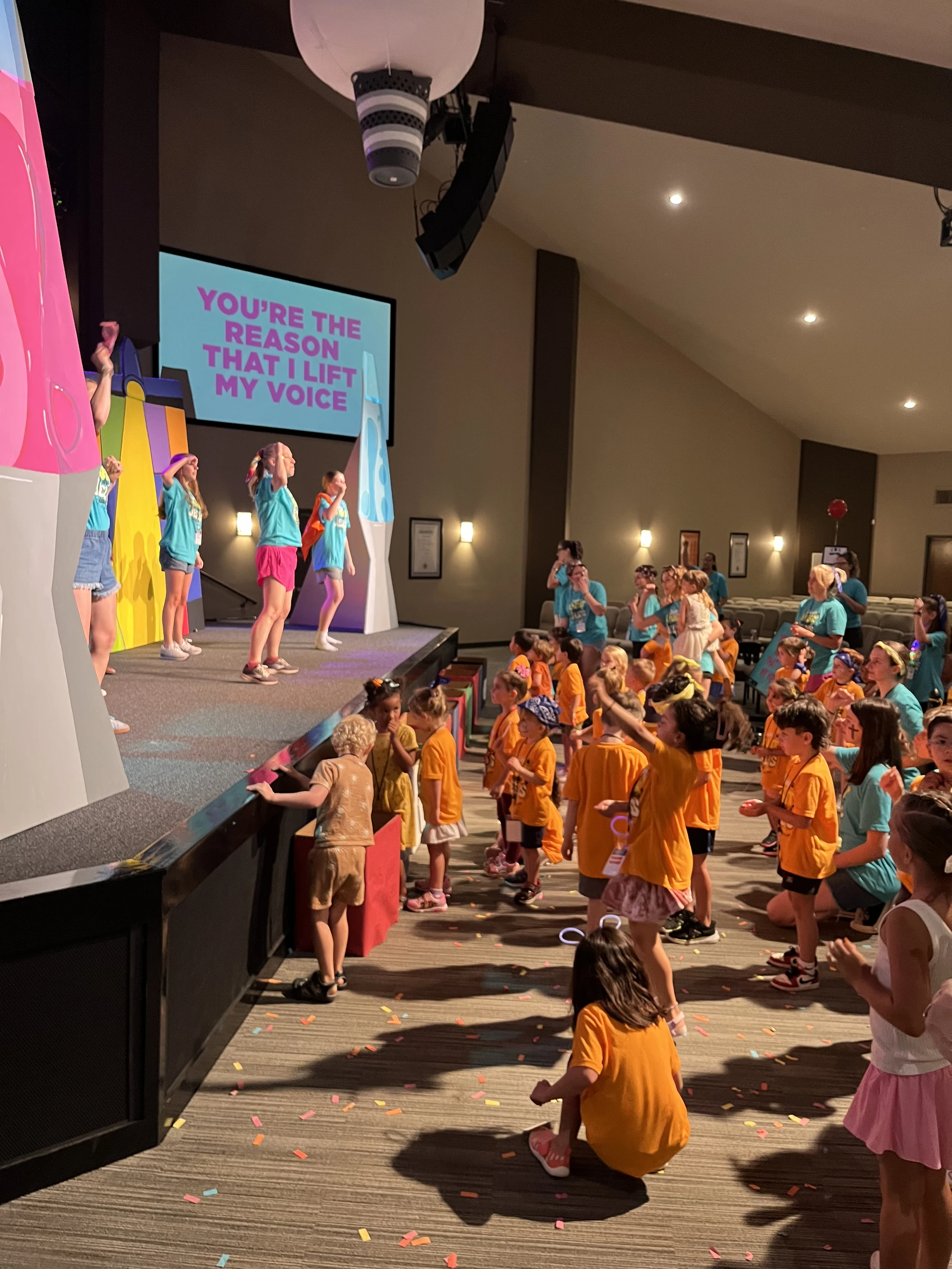 A large group of children in orange shirts watch other kids in blue shirts sing and dance on a brightly lit stage with a large screen displaying lyrics.