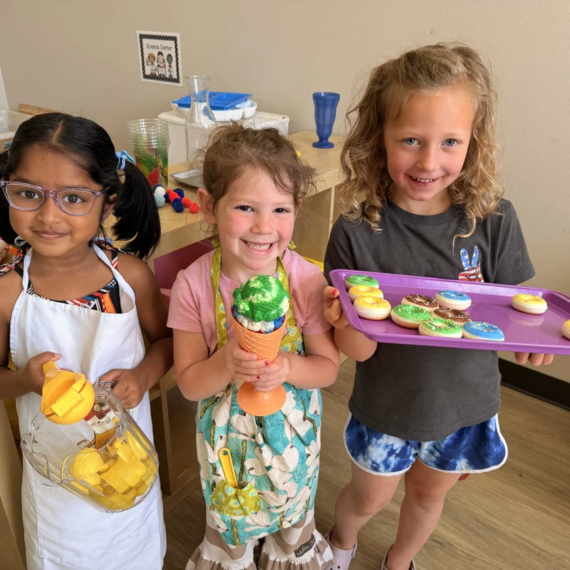 Three smiling young girls play with toy food. One holds a pitcher, another an ice cream cone, and the third holds a tray of toy donuts in a classroom.