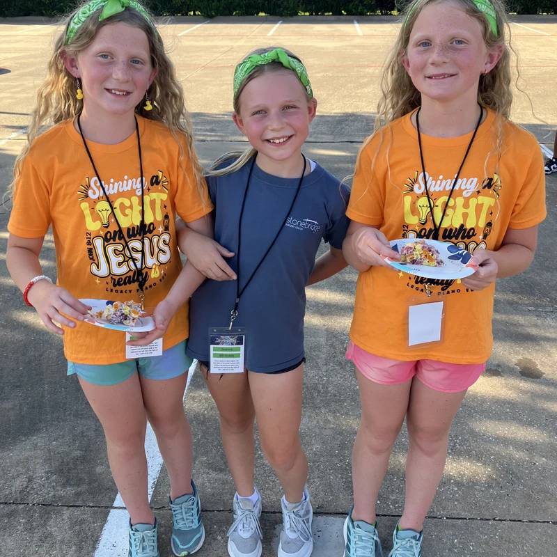 Three young girls stand together smiling in a parking lot. Two wear matching orange t-shirts and green bandanas, while holding paper plates with snacks.