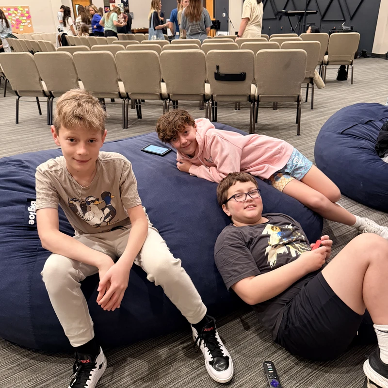 Three young boys smile while relaxing on a large blue beanbag chair in an auditorium. In the background, other people stand and talk near rows of chairs.