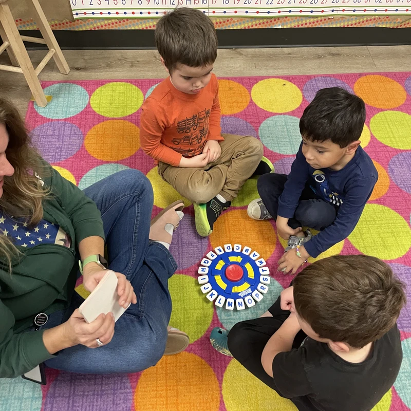 A teacher sits cross-legged on a colorful polka-dot rug with three young boys, playing a circular alphabet word game in a classroom.