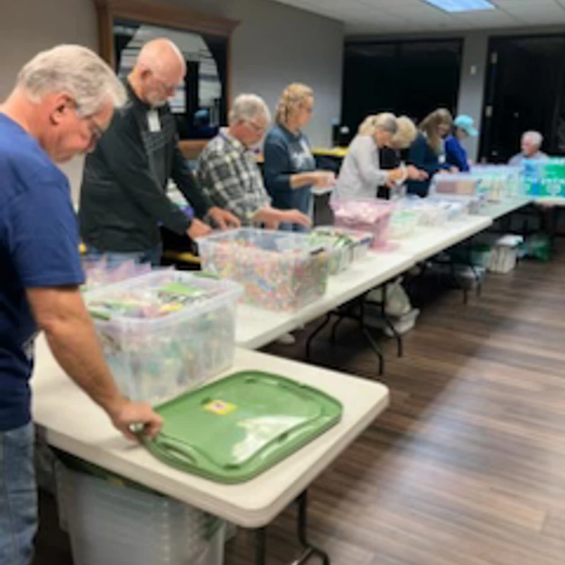 A group of people stand in a line along a long table, assembling packages from clear bins filled with small items.