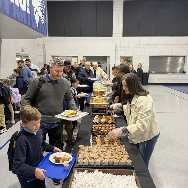 A long line of adults and children get food from a breakfast buffet table set up in a school gymnasium during a community event.