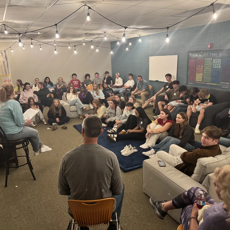 A large group of young people sit on couches and the floor, attentively listening to a woman speaking from a stool in a casually decorated room.