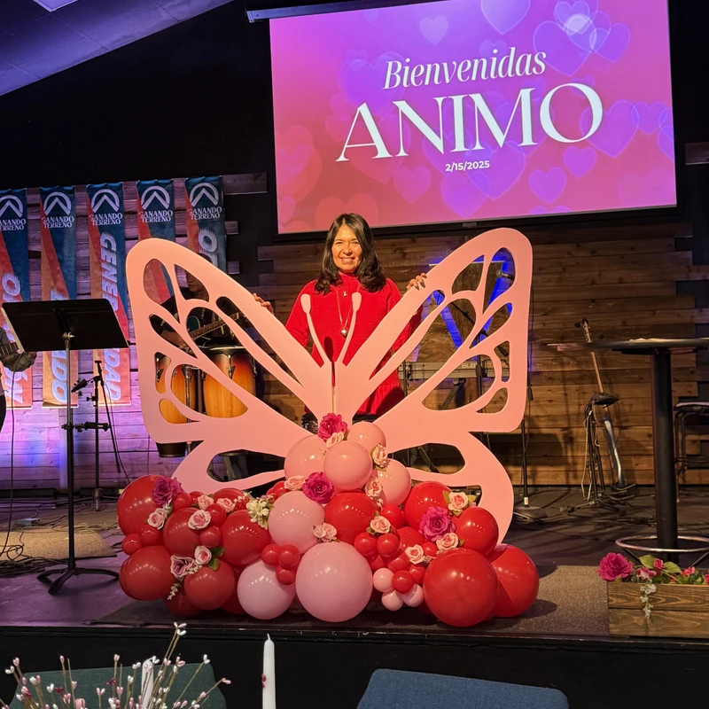 A woman in a red sweater smiles as she stands on a stage behind a large pink butterfly cutout decorated with red and pink balloons for an event.