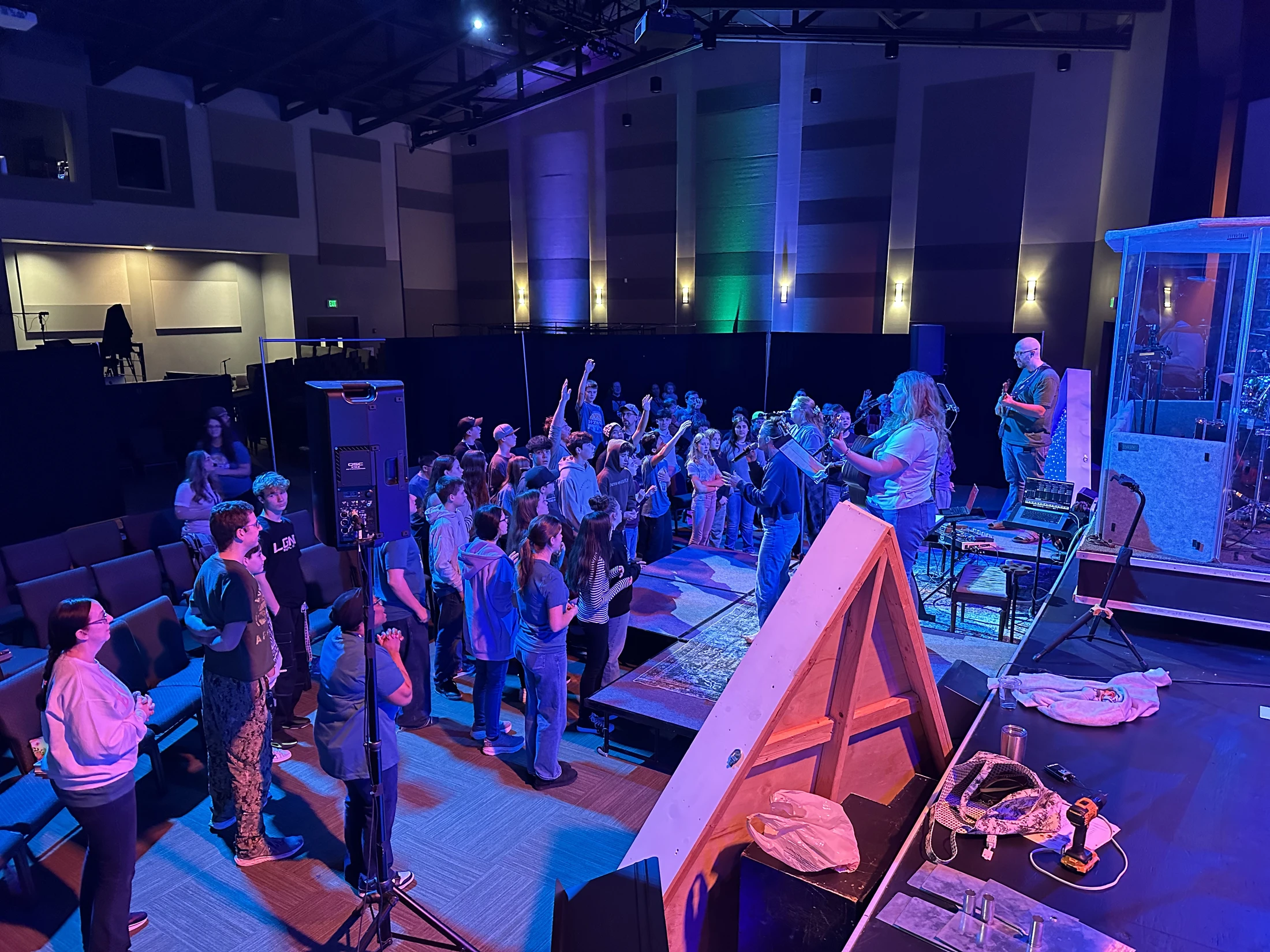 A crowd of young people stands before a stage in an auditorium, many with hands raised in worship as a band plays under blue and purple lights.