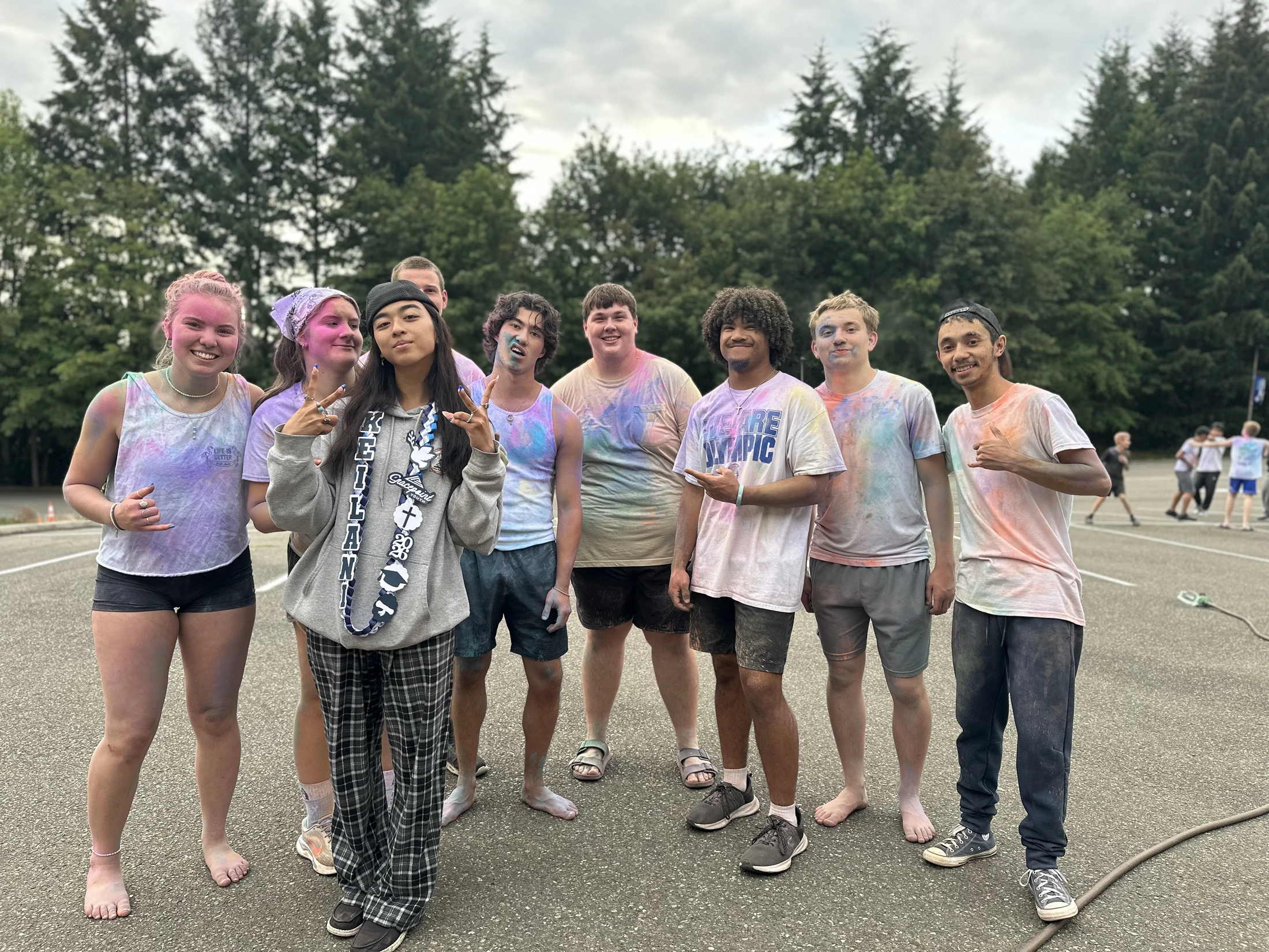 A diverse group of nine teenagers smile for a photo in a parking lot, their clothes and skin covered in brightly colored powder after a fun color fight.