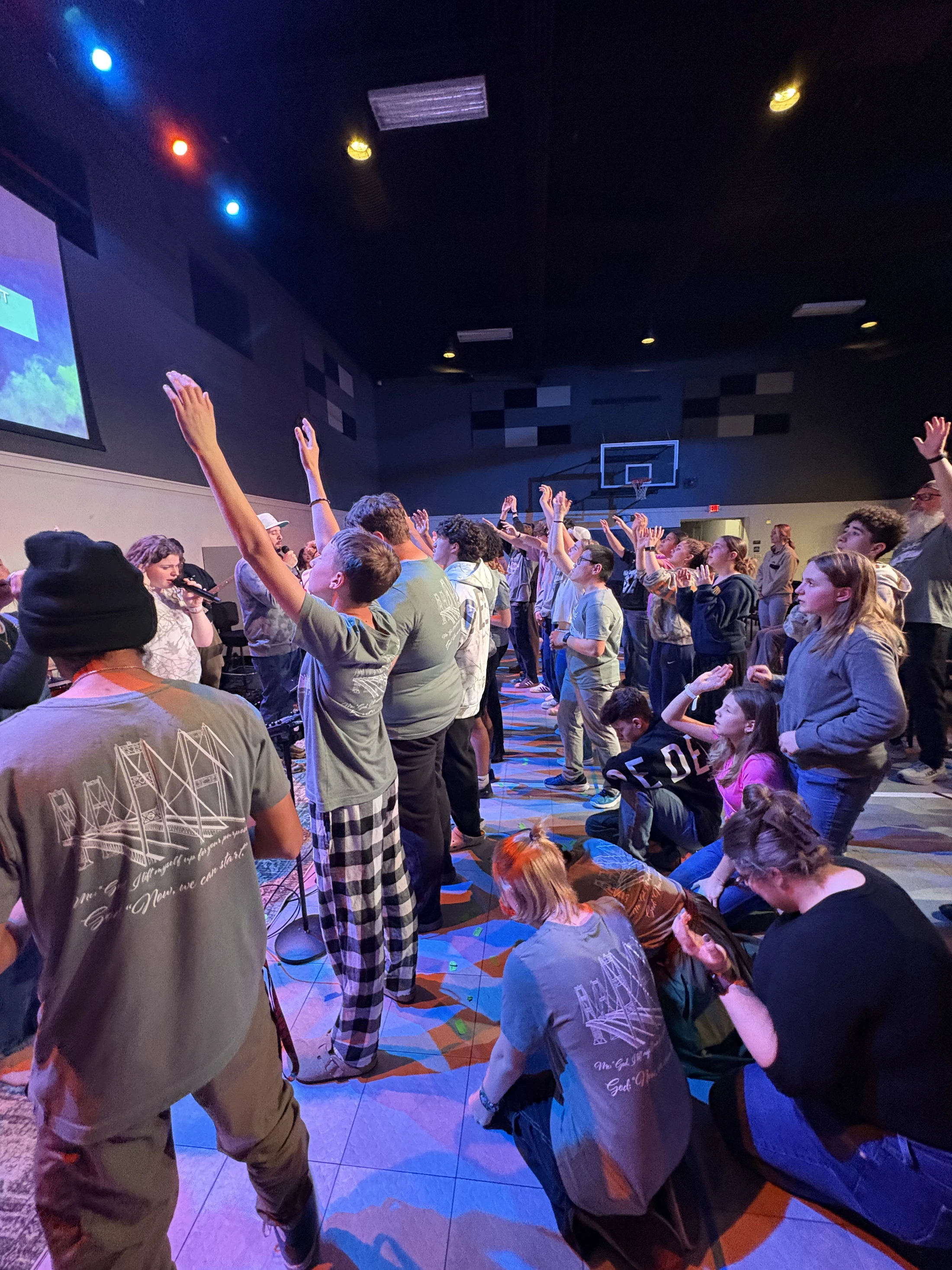 A large group of young people worship together in a dimly lit room. Many have their hands raised as they stand and sit facing a stage with colorful lights.