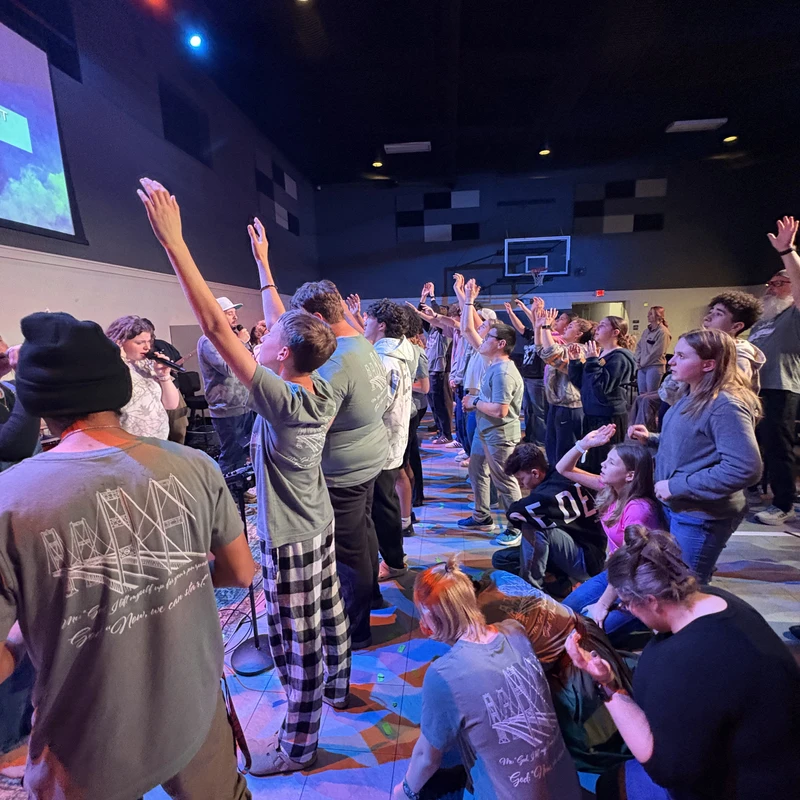 A large group of young people worship together in a dimly lit room. Many have their hands raised as they stand and sit facing a stage with colorful lights.