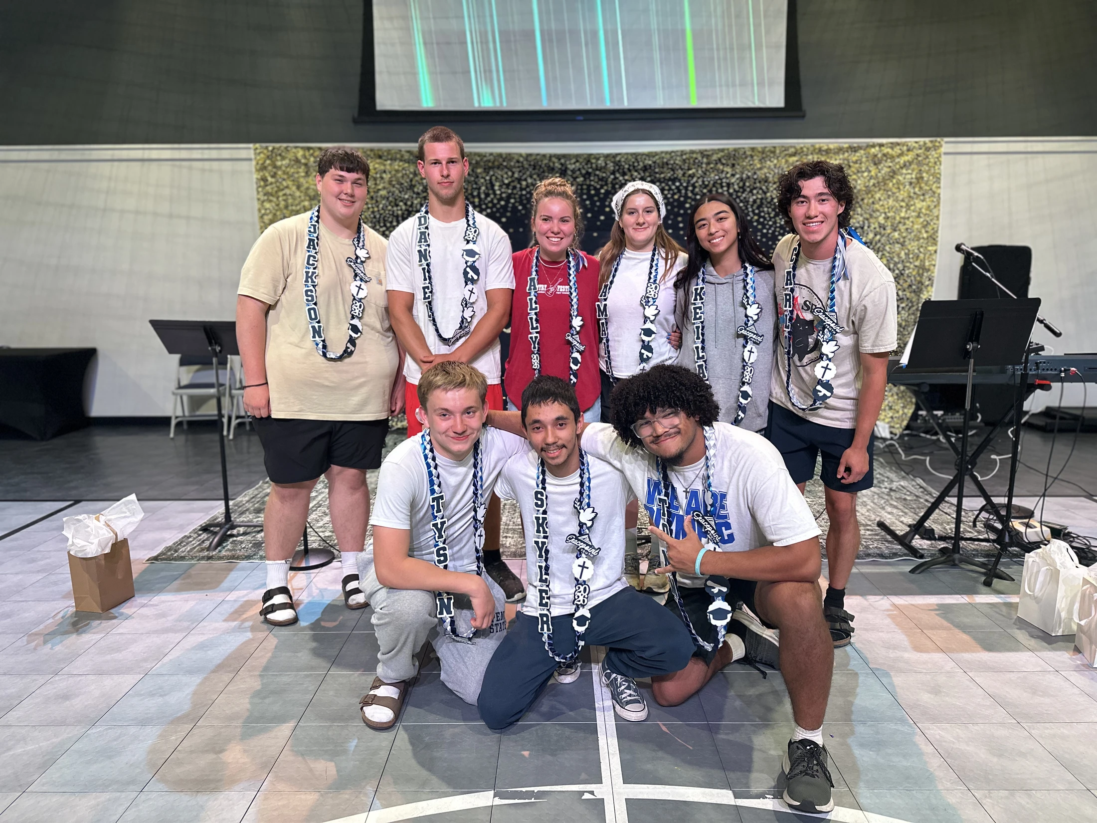 A group of nine smiling young adults pose for a photo on a stage. They are all wearing personalized leis, suggesting a graduation or celebration.