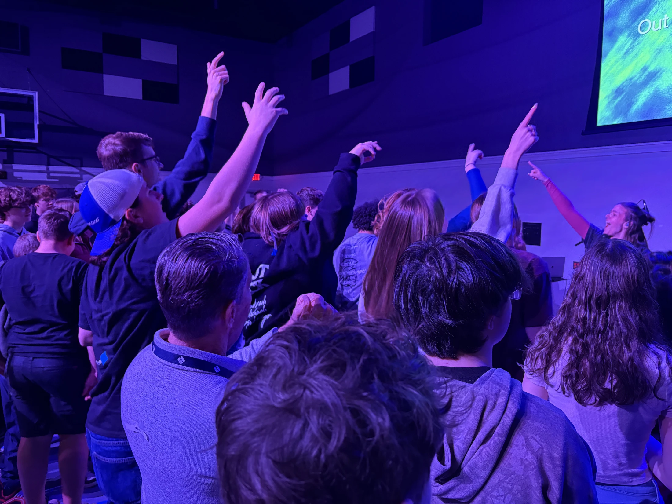 A crowd of young people, seen from behind, with their hands raised in a room filled with purple light during a concert or worship service.