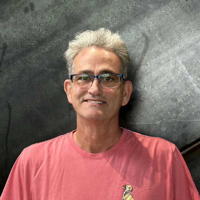 A headshot of a smiling man with short grey hair and blue-framed glasses, wearing a pink t-shirt while standing in front of a dark grey textured wall.