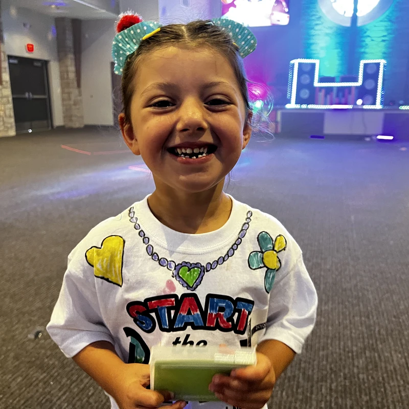 A young girl with a big smile wearing a decorated t-shirt and a cupcake liner hair bow. She is standing indoors with a colorful, lit stage in the background.
