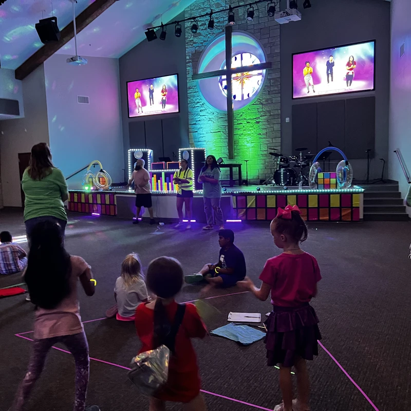 Children and adults dance and watch screens at a glow-in-the-dark party inside a church, with a brightly lit stage and cross in the background.