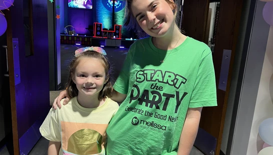 Two smiling girls, an older one in a green 'Start the Party' t-shirt with her arm around a younger girl, stand in a room decorated for a party.